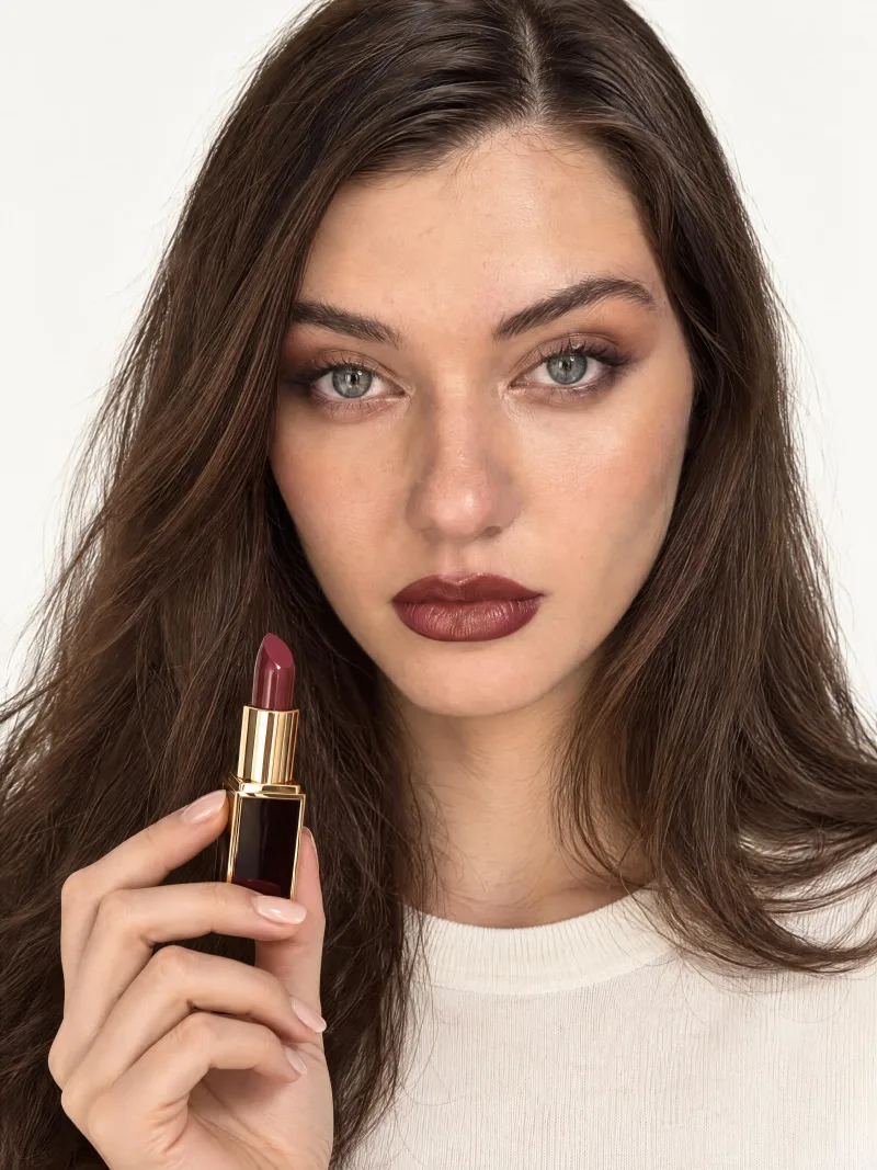 Dana facing the camera in full glam makeup, holding a burgundy lipstick with a gold casing